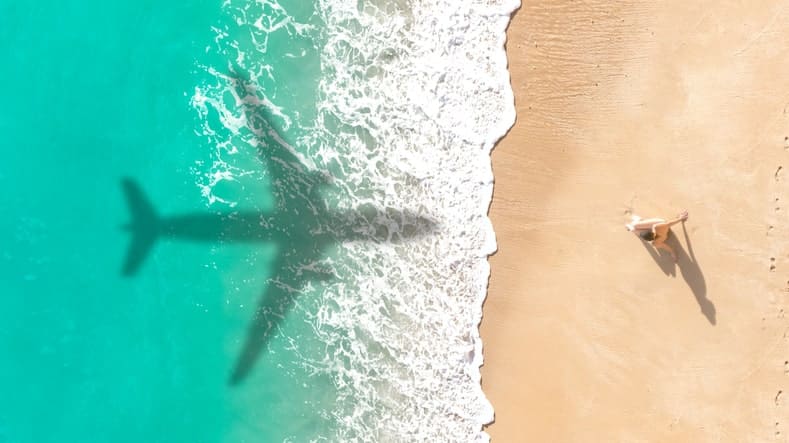 Aerial view of a beach with a vast airplane shadow cast over the water and sand. A person lies on the shore, perhaps dreaming of distant industries beyond the horizon.