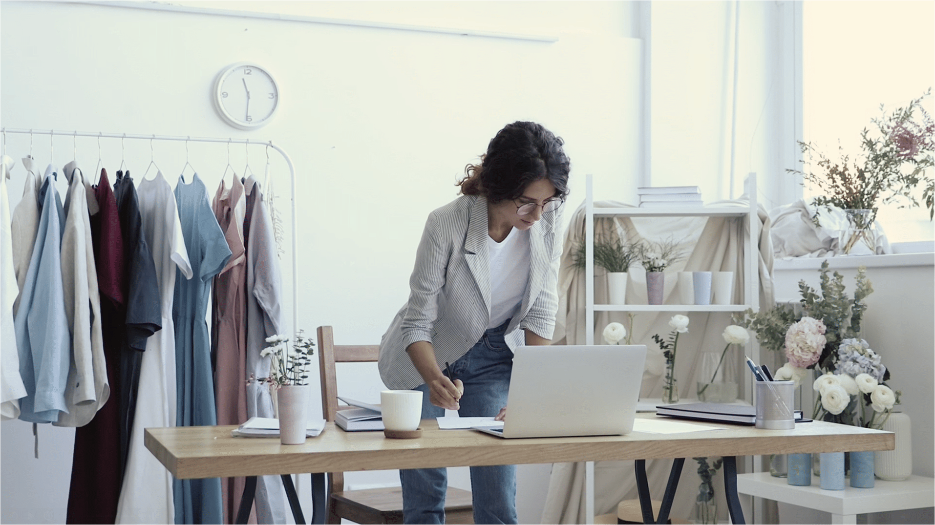 A woman wearing glasses stands at a desk, working on a laptop. The room has a clothing rack, plants, and a clock on the wall.
